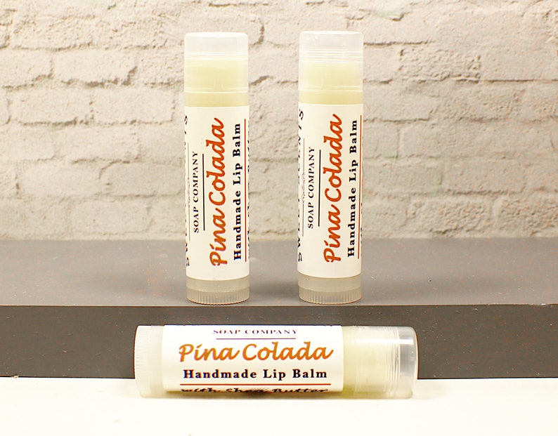K&ouml;nnte beinhalten: Drei wei&szlig;e Lippenbalsam-Tuben mit einem wei&szlig;en Etikett, auf dem "SOAP COMPANY" und "Pina Colada Handmade Lip Balm" in orangefarbenem und schwarzem Text steht.