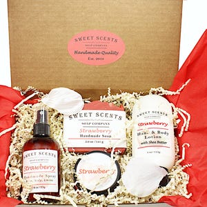 May include: A gift box filled with strawberry-scented bath and body products. The box contains a bar of soap, a bottle of body spray, a jar of body butter, and a bottle of hand and body lotion. All products are labeled "Strawberry" and "Sweet Scents Soap Company".