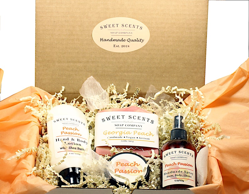 May include: A gift box filled with peach-scented bath and body products from Sweet Scents Soap Company. The box contains a bottle of hand and body lotion, a bar of soap, a jar of body butter, and a bottle of room spray. All products are labeled with the scent "Peach Passion" or "Georgia Peach".