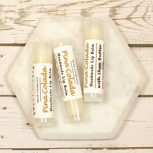 K&ouml;nnte beinhalten: Drei wei&szlig;e Lippenbalsam-Tuben mit einem wei&szlig;en Etikett, auf dem "Pina Colada Handgemachter Lippenbalsam mit Sheabutter" steht, auf einem wei&szlig;en, sechseckigen Marmorteller. Der Lippenbalsam befindet sich auf einer wei&szlig;en Holzoberfl&auml;che.