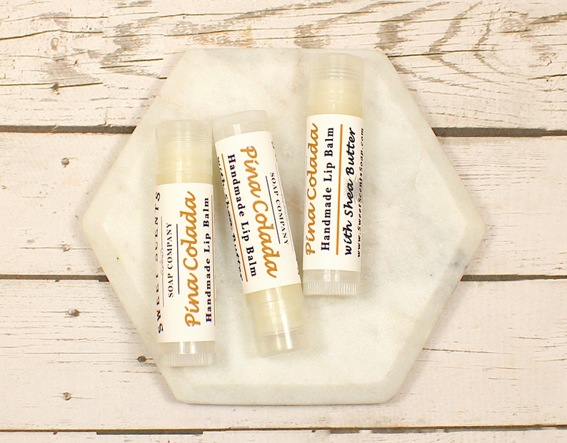 K&ouml;nnte beinhalten: Drei wei&szlig;e Lippenbalsam-Tuben mit einem wei&szlig;en Etikett, auf dem "Pina Colada Handgemachter Lippenbalsam mit Sheabutter" steht, auf einem wei&szlig;en, sechseckigen Marmorteller. Der Lippenbalsam befindet sich auf einer wei&szlig;en Holzoberfl&auml;che.