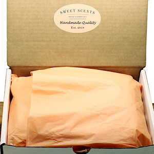May include: An open cardboard box with a white interior. The box is lined with peach-colored tissue paper. The box has a label on the top that reads "Sweet Scents Soap Company Handmade Quality Est. 2018".