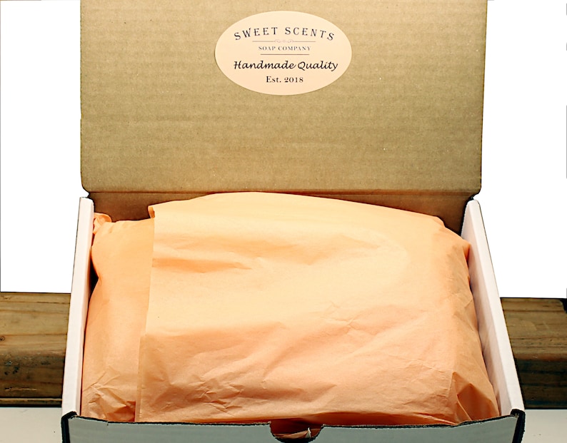 May include: An open cardboard box with a white interior. The box is lined with peach-colored tissue paper. The box has a label on the top that reads "Sweet Scents Soap Company Handmade Quality Est. 2018".
