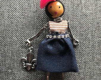 Op de afbeelding: Een zilveren ketting met een poppenhanger. De pop draagt een roze hoed, een gestreept shirt, een blauwe rok en een zilveren fleur-de-lis-hanger.