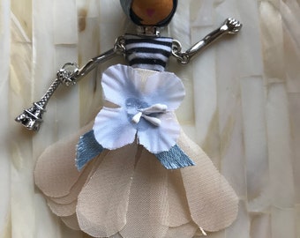 Op de afbeelding: Een witte pop met een blauw-wit gestreepte jurk, een witte bloem op haar hoofd en een zilveren Eiffeltoren-bedel. De pop draagt zilveren schoenen en een zilveren ketting.