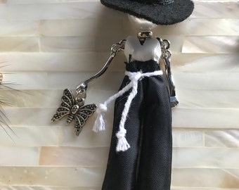 Op de afbeelding: Een zwart-witte poppenhanger met een zwarte hoed, een wit topje en een zwarte broek. De pop houdt een zilveren vlinderhanger vast.