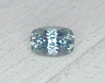 May include: A cushion-cut gemstone with a pale blue and green hue. The gem has a faceted surface that reflects light, creating a sparkling effect. The stone is set against a textured, light-colored background.
