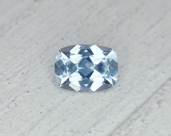 May include: A light blue cushion-cut gemstone with facets. The gemstone is set against a light gray background. The gem's shape is rectangular with rounded corners, and it reflects light, creating a sparkling effect.