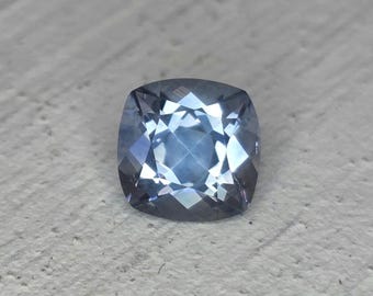 May include: A faceted, cushion-cut gemstone with a cool blue hue. The gem's surface reflects light, creating a sparkling effect. The stone is set against a neutral background, highlighting its color and cut. Ideal for jewelry.