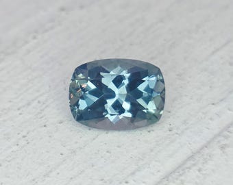 May include: A cushion-cut gemstone with a mix of blue and teal hues. The facets of the gem reflect light, creating a sparkling effect. The gemstone is set against a white surface.