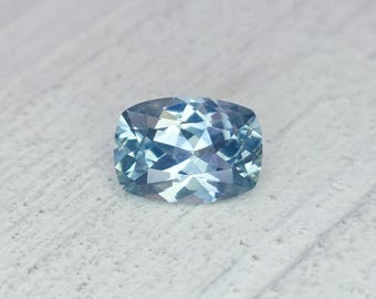 May include: A faceted, light blue gemstone with a cushion cut. The gem has a rectangular shape with rounded corners and a brilliant, reflective surface. The color is a pale, icy blue, with hints of lighter and darker shades within the facets.