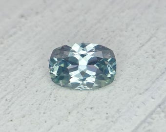 May include: A faceted, cushion-cut gemstone with a blend of blue and green hues. The gem is set against a textured, off-white surface, showcasing its multiple facets and internal reflections. The overall shape is a rounded rectangle.
