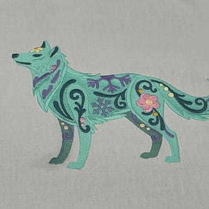 May include: An embroidered teal wolf design with floral and snowflake accents in purple, pink, and yellow. The wolf is standing, with a detailed pattern of swirls and dots. The design is on a light gray fabric.
