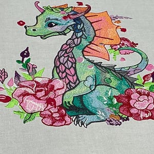 May include: Embroidered image of a whimsical dragon in shades of green, blue, and pink, surrounded by pink and red flowers. The dragon has orange wings and a decorative floral crown. The design is set against a light gray background.