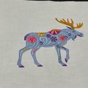 May include: Embroidered moose design on a white fabric. The moose is light blue with floral and snowflake patterns in pink, yellow, and purple. The antlers are orange. The design is likely for a craft project or home decor.