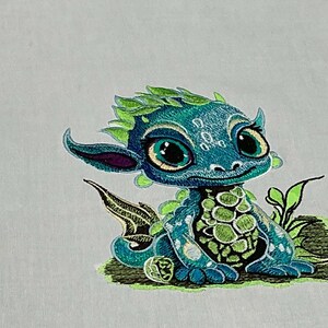 May include: Embroidered teal and green dragon with large eyes and a friendly expression. The dragon has green leafy accents on its head and tail, and a light green belly. The embroidery is on a light gray background.