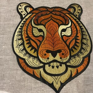 May include: Embroidered tiger head patch in orange, gold, and black. The tiger's face is intricately detailed with geometric patterns. The patch is likely for clothing or accessories.