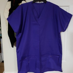 Puede incluir: Una bata quirúrgica de color morado con cuello en V y dos bolsillos. Los bolsillos tienen alfileres visibles.