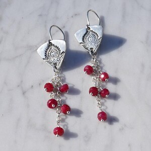 May include: A pair of silver dangle earrings with a triangular top featuring an etched design. Each earring has a chain of small, round, red beads. The earrings are displayed on a white marble surface.
