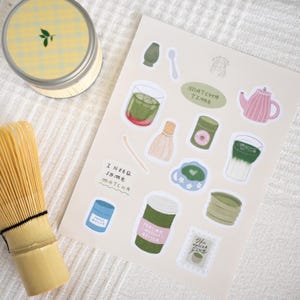 May include: A collection of matcha-themed stickers and a bamboo whisk. The stickers feature illustrations of matcha drinks, tea sets, and text that reads "Matcha Time" and "I need some matcha". A yellow and white tin is in the background.