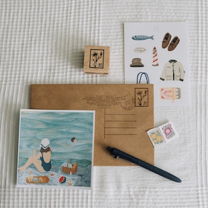 May include: A selection of stationery items, including a postcard with a beach scene, a brown envelope, a wooden stamp, and various stickers. The postcard depicts a woman at the beach. Stickers include a lighthouse, shoes, and a jacket.