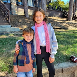May include: Two children wearing handmade scarves. One child wears a navy blue scarf with a brown shirt and a plaid jacket. The other child wears a pink scarf, a white shirt, a gray jacket, and black pants. The scarves appear to be knitted.