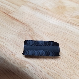May include: A small, dark gray, rectangular object with a textured surface. The object is lying on a light brown wooden surface. The object appears to be a piece of material or a small sculpture.