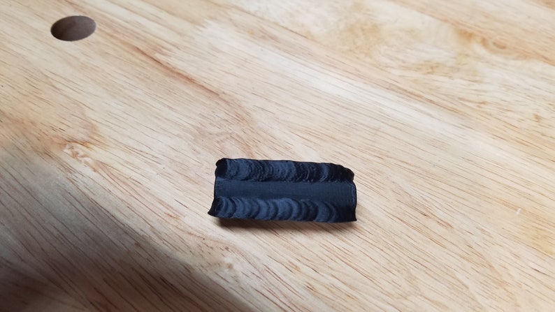 May include: A small, dark gray, rectangular object with a textured surface. The object is lying on a light brown wooden surface. The object appears to be a piece of material or a small sculpture.