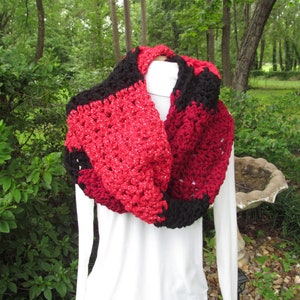 May include: A chunky, handmade infinity scarf in a bold color combination. The scarf features alternating sections of red and black yarn, with a speckled, glittery effect in the red sections. The scarf is displayed on a white form against a natural background.
