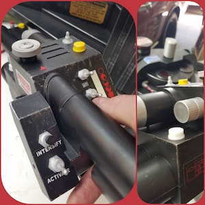 May include: A black prop device with various knobs, switches, and a cylindrical barrel. The device has a weathered appearance, with labels reading "INTENSIFY" and "ACTIVATE". Red and yellow accents are visible.