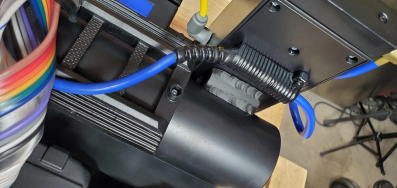 Może przedstawiać: Czarny cylindryczny element z żebrowanym metalowym wspornikiem montażowym, wyposażony w niebieski przew&oacute;d pneumatyczny połączony za pomocą czarnej elastycznej odgiętki. Po lewej stronie widoczna jest wiązka wielokolorowych przewod&oacute;w oraz ż&oacute;łty wąż.