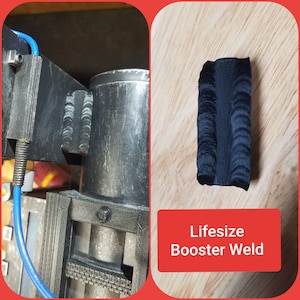 May include: A black, textured, rectangular Lifesize Booster Weld prop, likely for cosplay or display. The prop is on a wooden surface. The image also shows a portion of a larger, metallic prop with a blue cable.