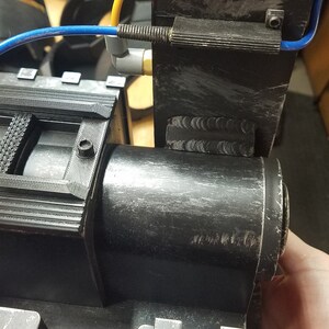May include: A close-up of a black, industrial-looking device with a cylindrical component and a rectangular section. A blue tube and other components are attached. The device appears to be made of plastic or a similar material.