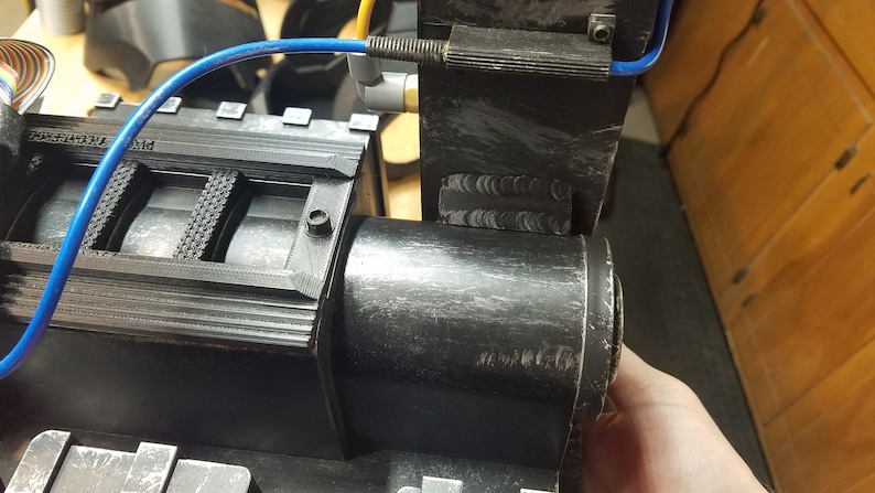 May include: A close-up of a black, industrial-looking device with a cylindrical component and a rectangular section. A blue tube and other components are attached. The device appears to be made of plastic or a similar material.