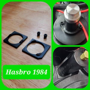 May include: Two black square plastic plates with circular cutouts, two small black cylindrical components, and a metallic knob assembly. The text Hasbro 1984 is visible. These components are shown in various orientations and close-up configurations.
