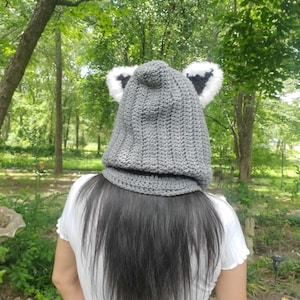 Puede incluir: Un gorro de ganchillo gris con orejas adjuntas, con ribete de piel sint&eacute;tica blanca y negra. El gorro est&aacute; dise&ntilde;ado para cubrir la cabeza y el cuello, ofreciendo calidez y una est&eacute;tica l&uacute;dica. El gorro se muestra al aire libre.
