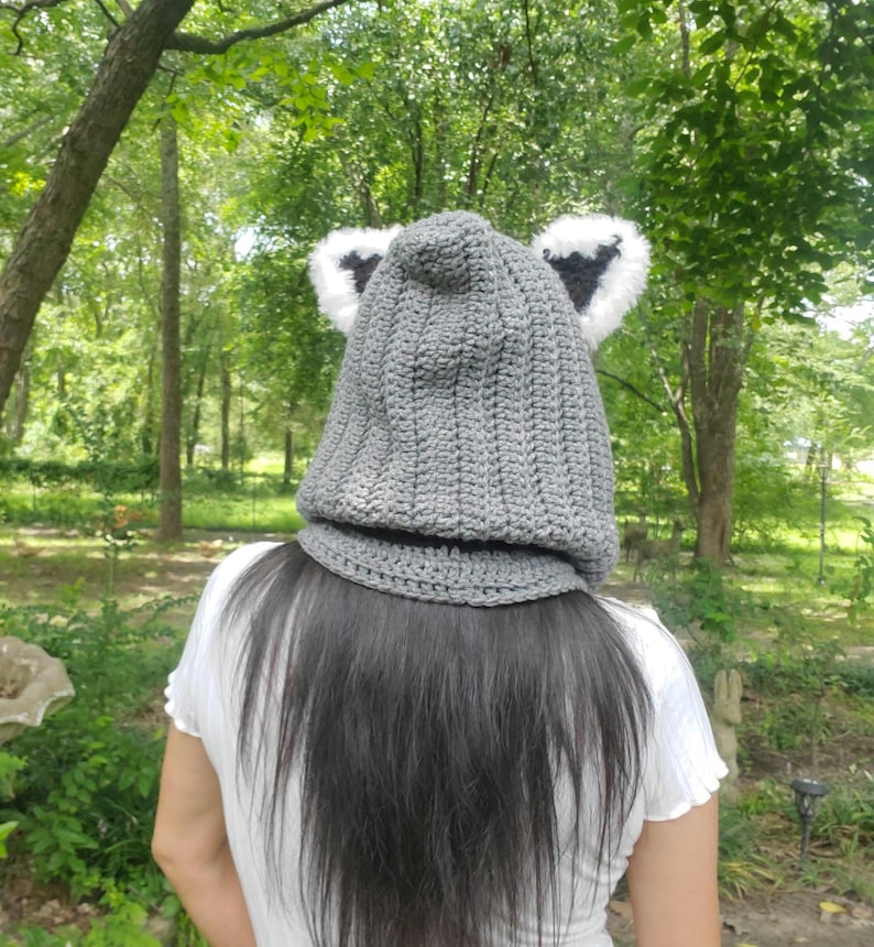 Puede incluir: Un gorro de ganchillo gris con orejas adjuntas, con ribete de piel sint&eacute;tica blanca y negra. El gorro est&aacute; dise&ntilde;ado para cubrir la cabeza y el cuello, ofreciendo calidez y una est&eacute;tica l&uacute;dica. El gorro se muestra al aire libre.