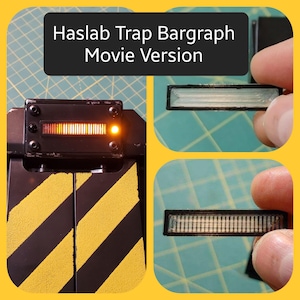 May include: A black and yellow prop with the text "Haslab Trap Bargraph Movie Version." The prop features a rectangular display with an illuminated bar graph and a yellow light. The trap has black and yellow diagonal stripes.