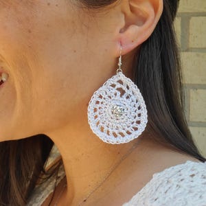 May include: White crochet earrings with a teardrop shape and a silver-colored hook. The earrings have a circular design with a sparkling gem in the center. The earrings are a handmade accessory.