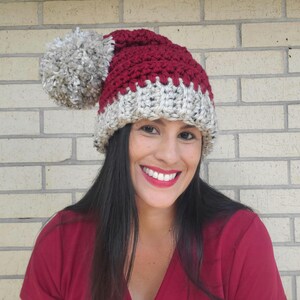 May include: A handmade, chunky-knit hat in burgundy, gray, and cream. The hat features a large, fluffy gray pom-pom and a cream-colored brim. The hat is a winter accessory.