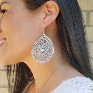 May include: A silver crochet teardrop earring with a central crystal. The earring is handmade with a delicate, open weave pattern. The earring hangs from a silver hook.