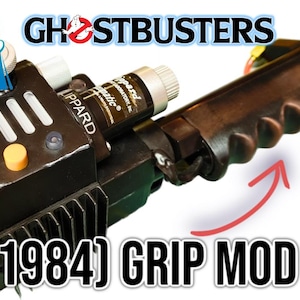 May include: A prop replica of a Ghostbusters proton pack with the Hasbro logo. The black and gray device features a handle, switches, and a gauge. The text "(1984) GRIP MOD" is visible.