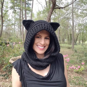 May include: A black crochet hood with cat ears and a matching cowl. The hood is worn over the head and shoulders. The background includes trees and pink flowers.