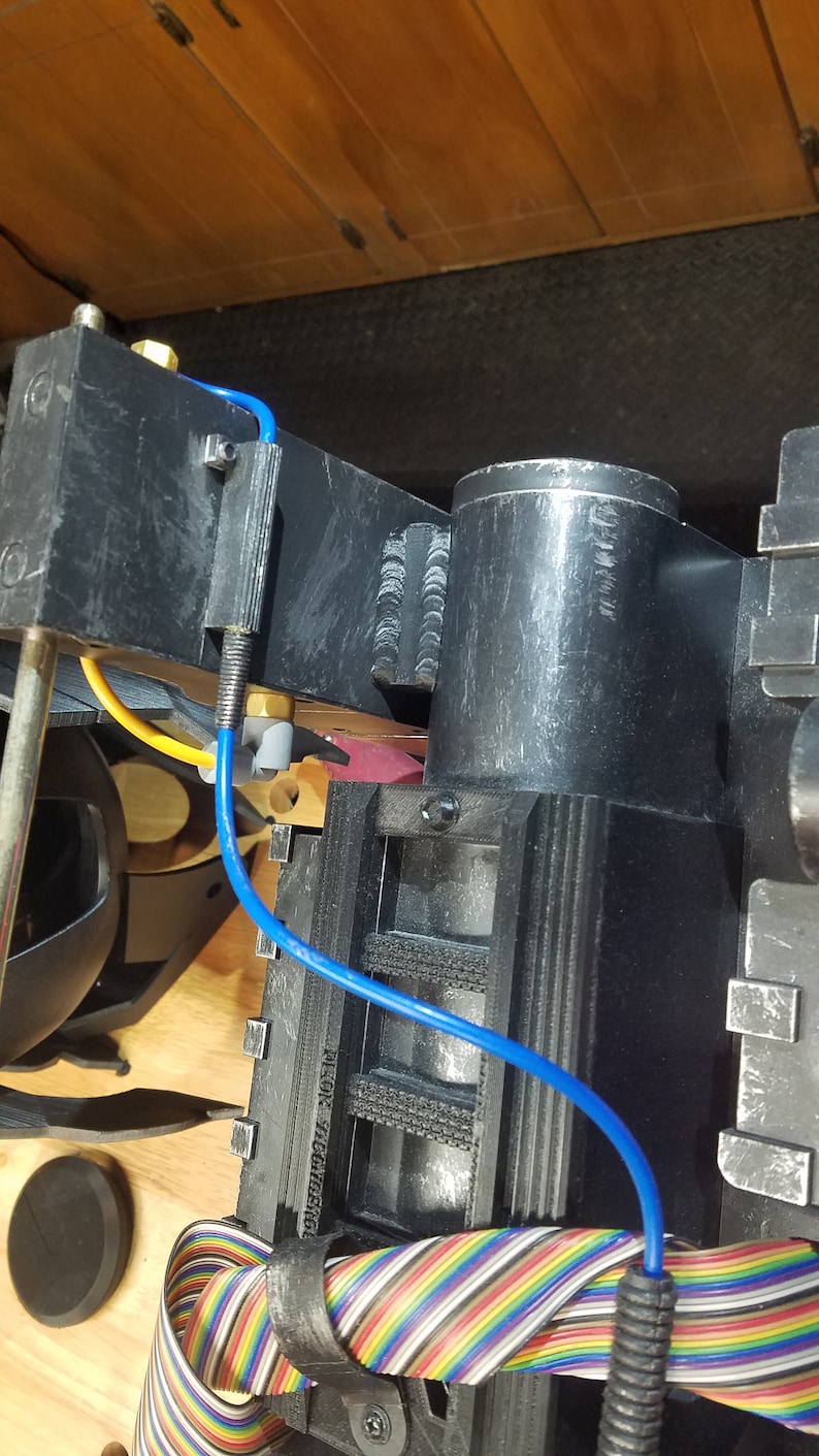 May include: A detailed view of a black prop device with a cylindrical component and various wires. A blue wire curves across the device, and a multi-colored ribbon cable is visible. The device is set against a wooden surface.