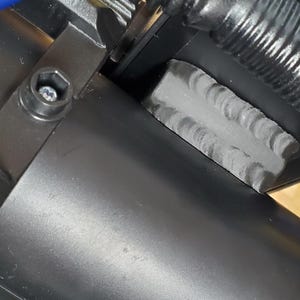 Może przedstawiać: Czarny zesp&oacute;ł mechaniczny z cylindryczną obudową, żebrowanym elementem metalowym i widoczną spoiną spawalniczą. Niebieski przew&oacute;d jest poprowadzony przez konstrukcję, kt&oacute;ra jest zabezpieczona śrubą sześciokątną i mechanizmem sprężyny zwojowej.