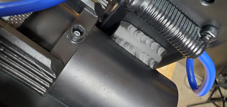 Może przedstawiać: Czarny zesp&oacute;ł mechaniczny z cylindryczną obudową, żebrowanym elementem metalowym i widoczną spoiną spawalniczą. Niebieski przew&oacute;d jest poprowadzony przez konstrukcję, kt&oacute;ra jest zabezpieczona śrubą sześciokątną i mechanizmem sprężyny zwojowej.