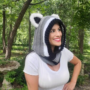 Puede incluir: Una capucha de ganchillo en gris, blanco y negro, dise&ntilde;ada para parecerse a un mapache. La capucha presenta orejas de piel sint&eacute;tica y un patr&oacute;n de rayas. La capucha la lleva una persona con una camisa blanca, sobre un fondo de &aacute;rboles y vegetaci&oacute;n.