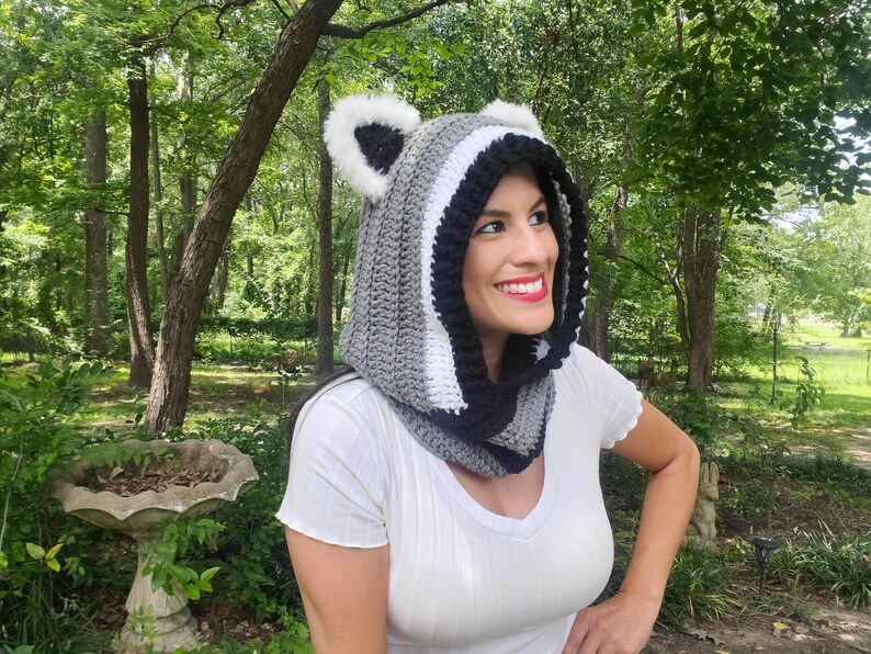 Puede incluir: Una capucha de ganchillo en gris, blanco y negro, dise&ntilde;ada para parecerse a un mapache. La capucha presenta orejas de piel sint&eacute;tica y un patr&oacute;n de rayas. La capucha la lleva una persona con una camisa blanca, sobre un fondo de &aacute;rboles y vegetaci&oacute;n.