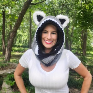 Puede incluir: Una capucha de mapache de ganchillo hecha a mano con una bufanda a juego. La capucha presenta un dise&ntilde;o a rayas negras y grises con orejas de piel sint&eacute;tica blanca. La bufanda es de rayas grises, negras y blancas. La capucha se usa alrededor del cuello y la cabeza.