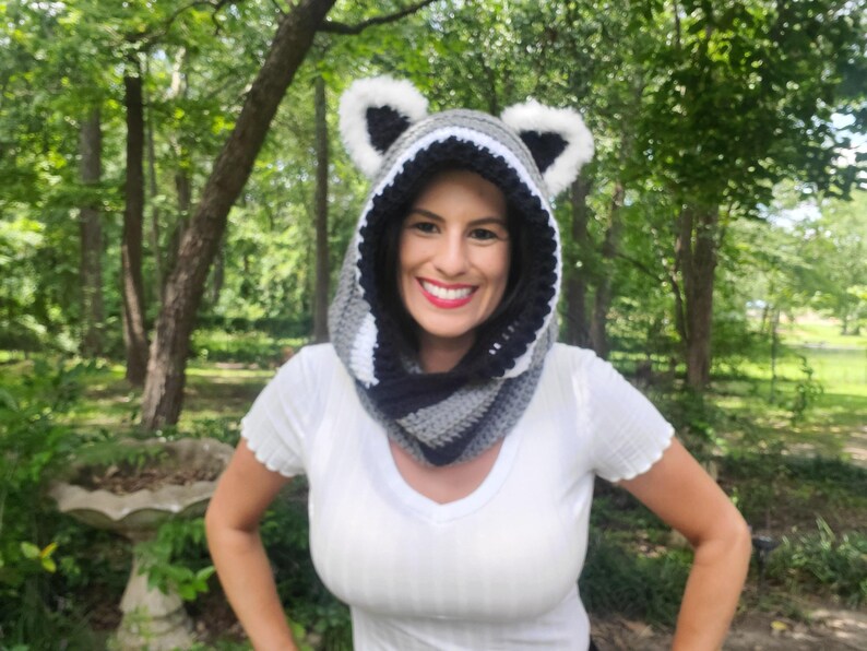 Puede incluir: Una capucha de mapache de ganchillo hecha a mano con una bufanda a juego. La capucha presenta un dise&ntilde;o a rayas negras y grises con orejas de piel sint&eacute;tica blanca. La bufanda es de rayas grises, negras y blancas. La capucha se usa alrededor del cuello y la cabeza.